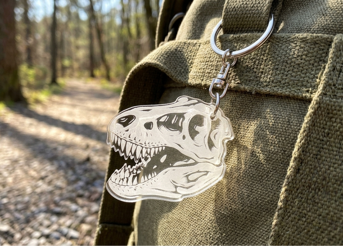 A clear acrylic keychain featuring a laser-cut T-rex skull fossil design, attached to a silver metal key ring and clipped to an olive green canvas backpack, with a blurred forest path in the background.