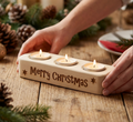 A wooden Christmas candle holder engraved with 'Merry Christmas' and stars, holding three lit tea light candles, shown on a rustic wooden table with hands holding it and holiday decor in the background.