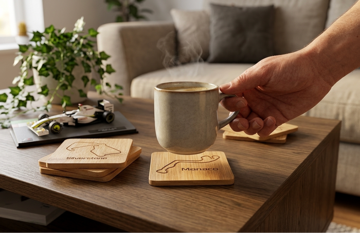 A person's hand holds a beige ceramic mug with steam rising, resting on a wooden race track coaster engraved with a Formula 1 track outline and the name 'Monaco'. The scene is set in a cozy living room on a wooden coffee table, alongside other engraved wooden coasters, an F1 model car, and a potted plant.