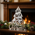 A silver mirror acrylic laser-cut Christmas tree shaped sign reading 'We Wish You a Merry Christmas' stands on an acrylic base on a wooden mantel, decorated with a festive garland and a warm fireplace in the background.