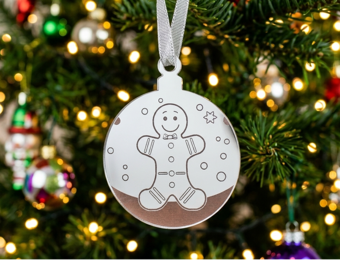 A round mirrored acrylic Christmas ornament with a gingerbread man outline and small snowflake details, hanging from a silver ribbon on a decorated Christmas tree with warm lights.