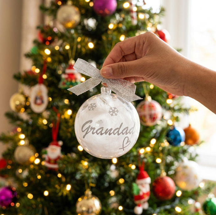 A hand holds a shatterproof bauble Christmas ornament filled with white feathers and 'Grandad' written in silver vinyl and a silver organza ribbon bow, set against a blurred decorated Christmas tree with warm lights.