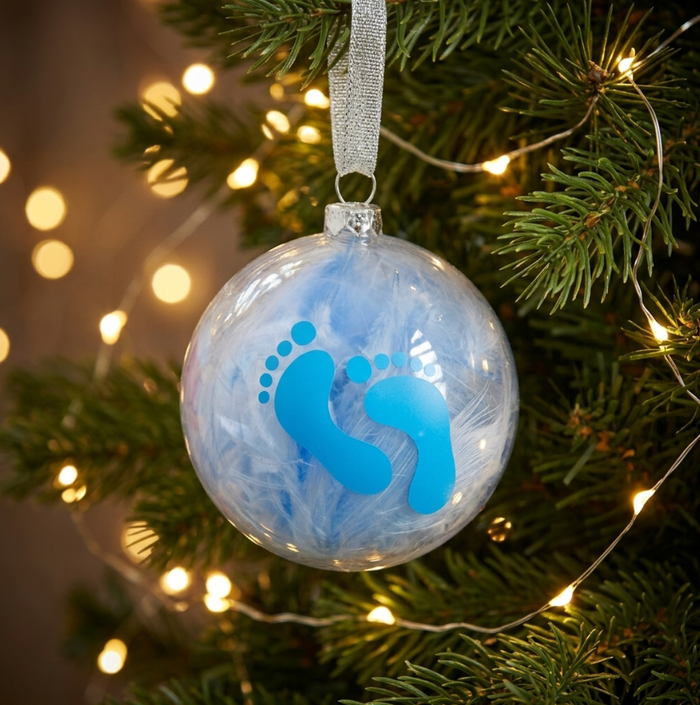 A clear round Christmas bauble ornament hanging on a pine tree, filled with blue feathers and featuring a bright blue baby footprint design. Warm string lights glow in the background.