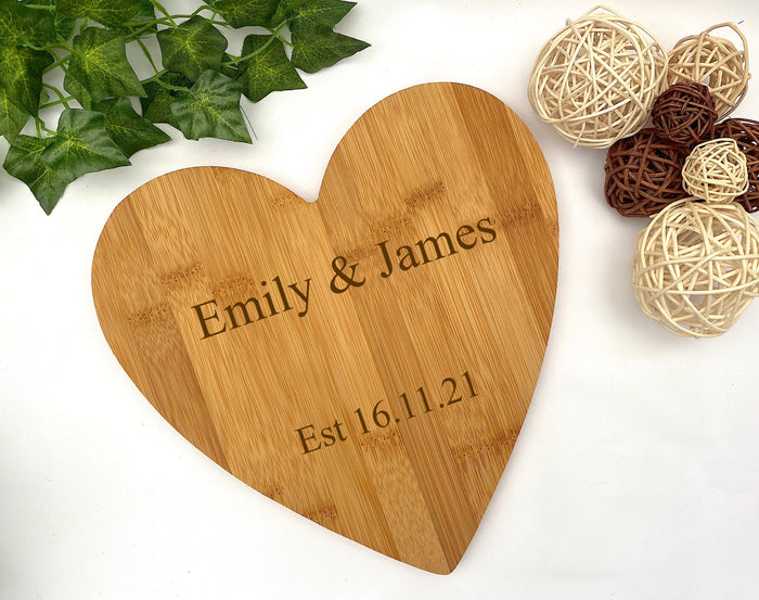 Wooden heart-shaped cutting board with engraved names and date, surrounded by decorative elements on a white background.