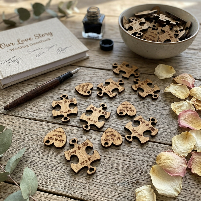 Personalised wooden wedding keepsake puzzle pieces and hearts arranged on a rustic wooden table, alongside a signed wedding notebook, a pen, a small ink bottle, a bowl filled with matching pieces, and scattered dried flower petals.