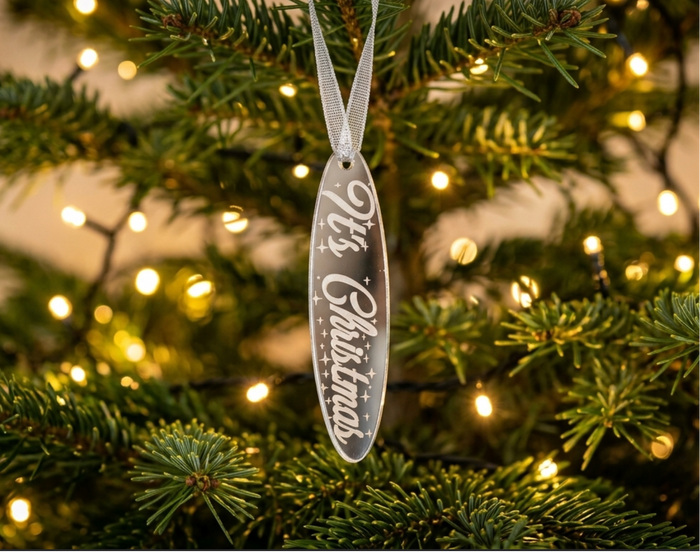 A silver mirror acrylic Christmas ornament with the words 'It's Christmas' engraved, hanging from a silver ribbon on a decorated Christmas tree with warm lights.