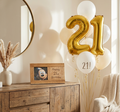 A personalised wooden photo frame to celebrate special occasions. A styled interior scene with a cluster of balloons including large metallic gold '21' balloons, white balloons, and one white balloon reading '21!'. A wooden cabinet, mirror, dried grass in a vase, and a knitted chair are visible in a warm room setting.