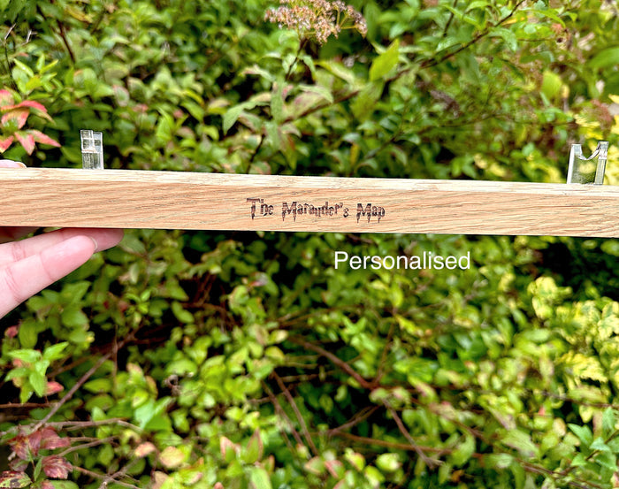 A hand holds a light-coloured wooden wand stand outdoors in front of a green leafy background. It has the words 'The Marauder's Map' engraved on it, and a white text overlay reads 'Personalised'. Harry Potter inspired wand stand.