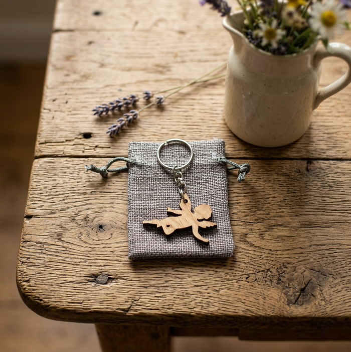 A small grey fabric drawstring pouch holding a silver keyring with a wooden angel-shaped charm, styled on a rustic wooden table with lavender sprigs and a ceramic pitcher of flowers in the background.