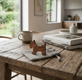 A rustic wooden table in a bright, modern kitchen setting, styled with a small wooden dog-shaped keyring on a grey cloth, a pair of glasses, and stacked books next to a ceramic mug.