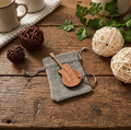 A wooden guitar-shaped keychain with a keyring, resting on a small grey fabric drawstring pouch. The scene is styled on a rustic wooden table with decorative yarn balls, green ivy, and ceramic mugs.