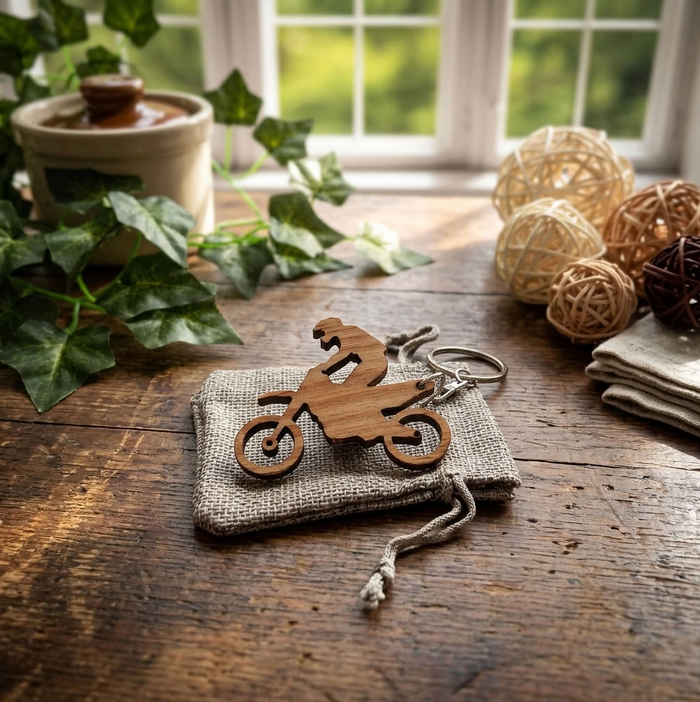 A wooden cutout keychain shaped like a dirt bike rider on a small burlap pouch, placed on a rustic wooden table with decorative plants, woven balls, and a window in the background.