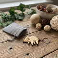 A wooden sloth keychain with a silver metal ring, displayed on a rustic wooden table alongside a small grey fabric pouch, decorative balls, and green leaves.