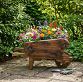 A rustic wooden wheelbarrow planter filled with a colorful assortment of petunias and marigolds, set on a stone patio in a lush garden with gardening tools nearby.