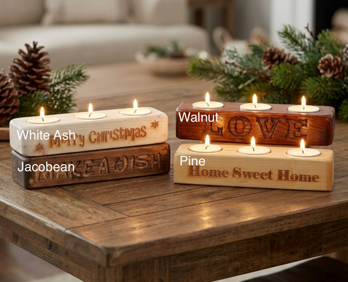 Four rectangular wooden candle holders with lit tea lights are arranged on a wooden table in a cozy indoor setting. The holders are shown in different wood finishes labeled in the image as White Ash, Jacobean, Walnut, and Pine.