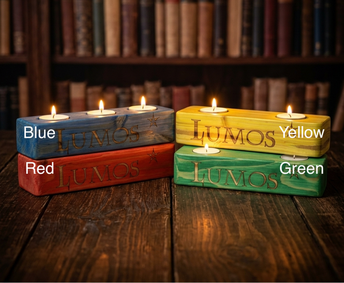 Four rectangular wooden candle holders with tea light candles, labelled Blue, Red, Yellow, and Green, arranged on a dark wooden table with a blurred bookshelf background. The words 'LUMOS' are engraved on each holder.